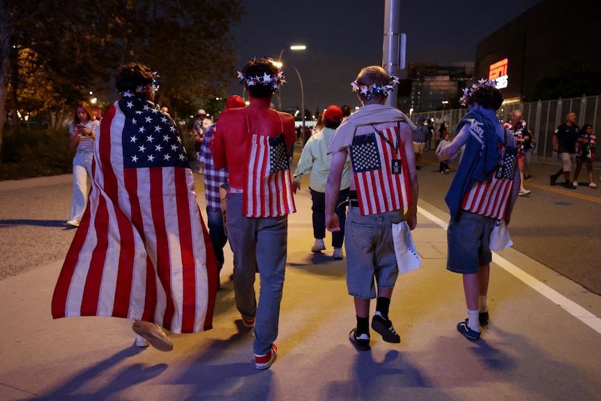 four people walking doan a street at night facing away, with US flags on their backs