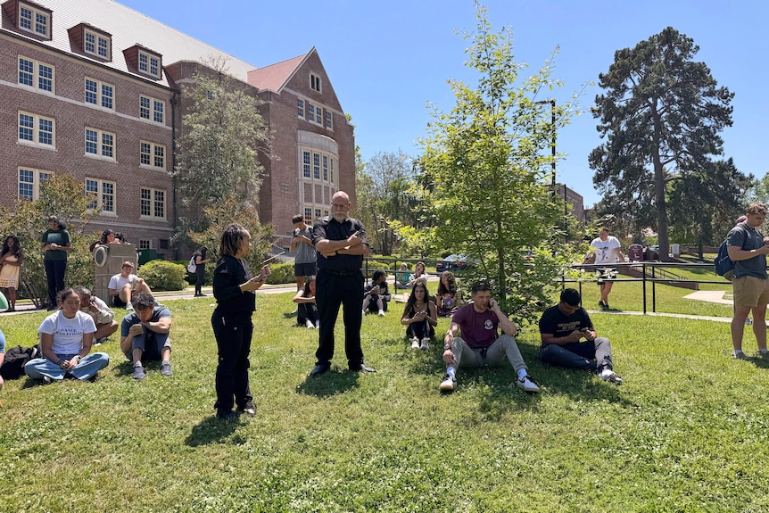 A crowd of people sit on the grass outside a brick building