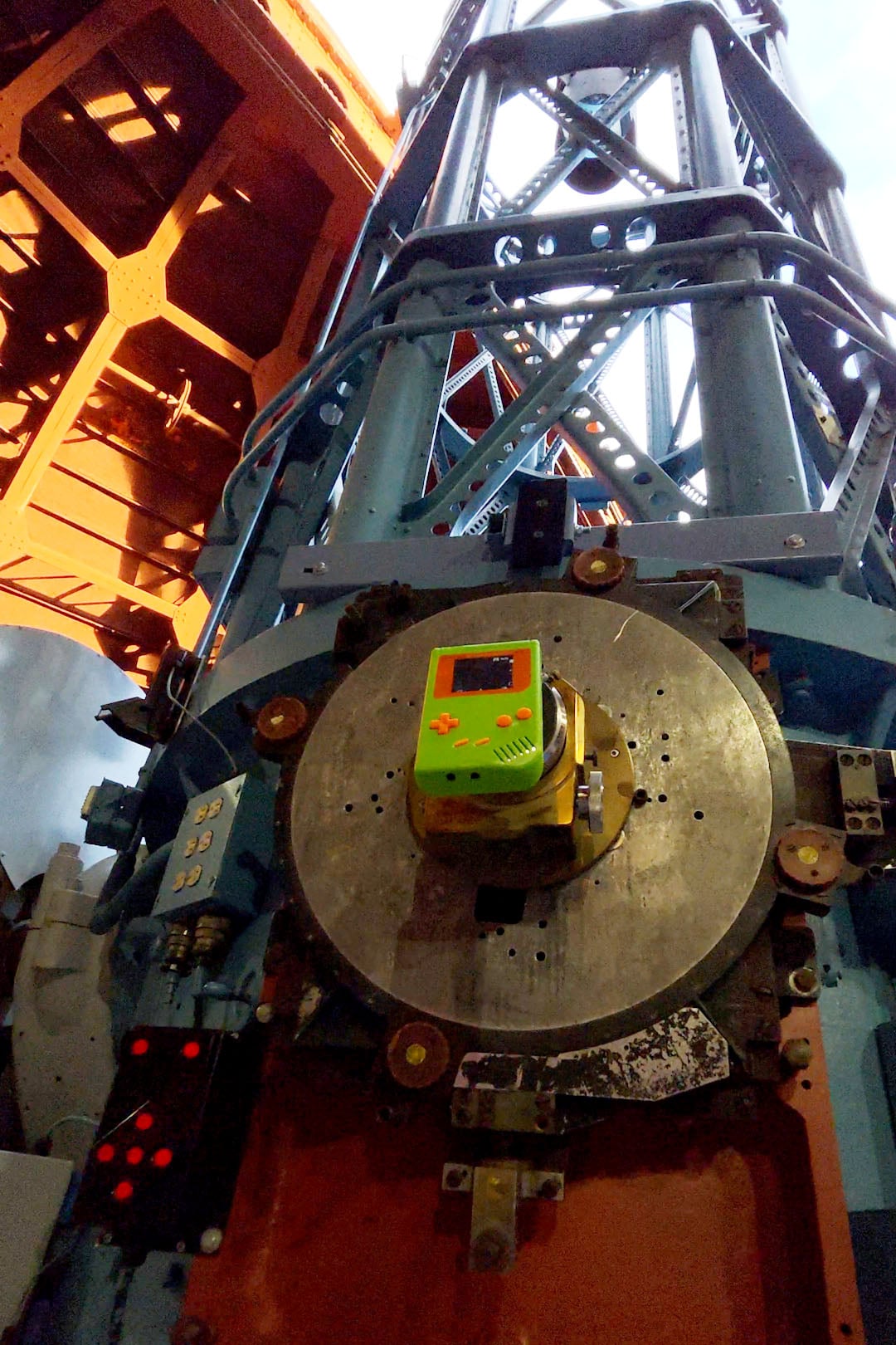 A green Game Boy is mounted on a large metal apparatus, possibly part of industrial or scientific equipment, with beams and structures extending upward in the background.