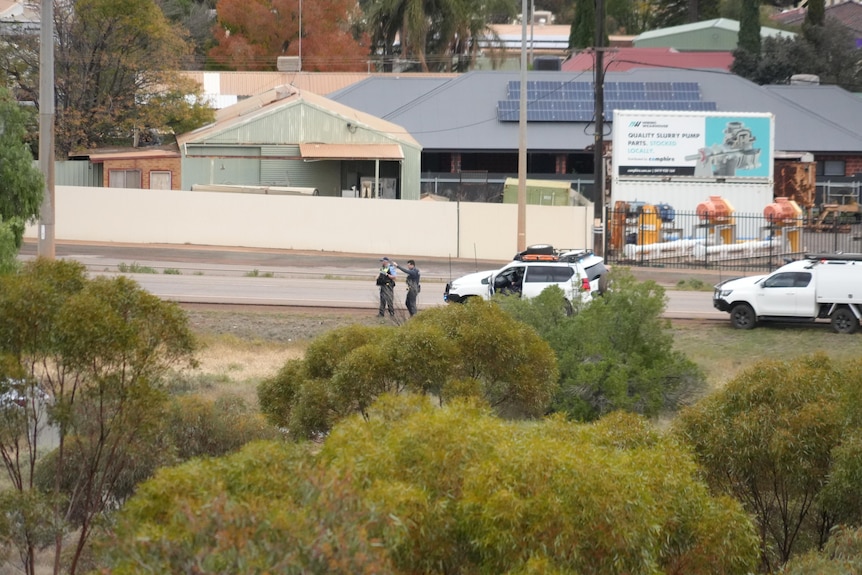 Two white cars parked at the side of a road with two men, one in a police uniform standing by them, bushland in the foreground