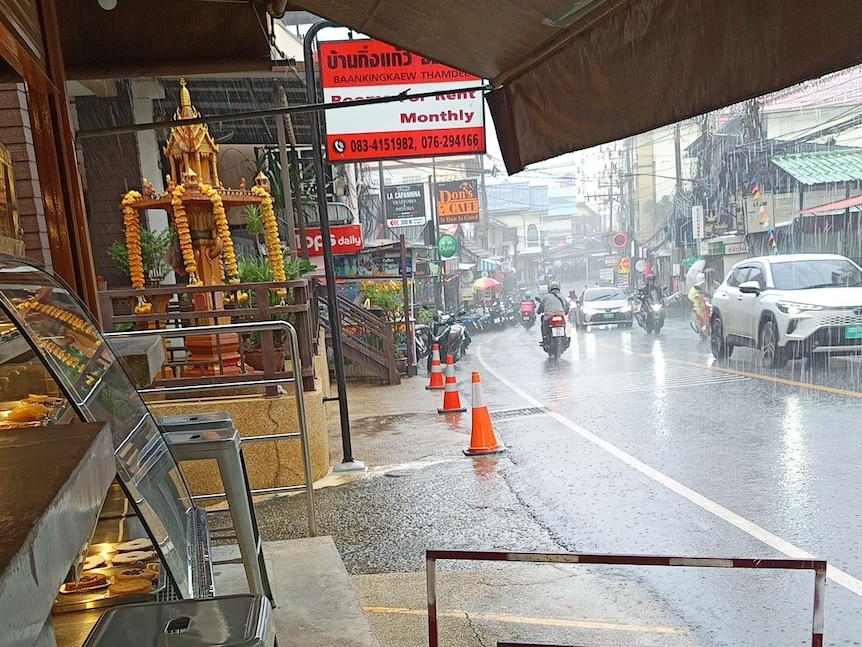 Rain pouring down on a street in Thailand with cars and motorbikes on the road.