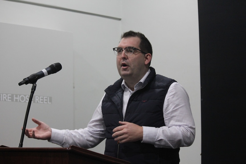 A dark-haired man in a dark vest stands at a lectern and speaks into a microphone.