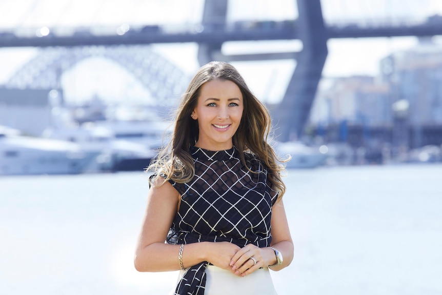 Tina has long hair and a dark shirt, and clasps her hands in front. She's in front of the Sydney Harbour Bridge.