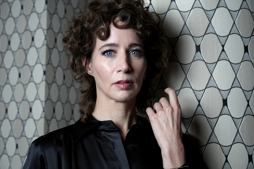 A close-up portrait of Miranda July, who has curled auburn hair and blue eyes and is standing against a tiled wall.