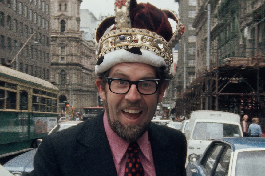 Rolf Harris as a young man standing on a busy city street wearing a large ornate crown