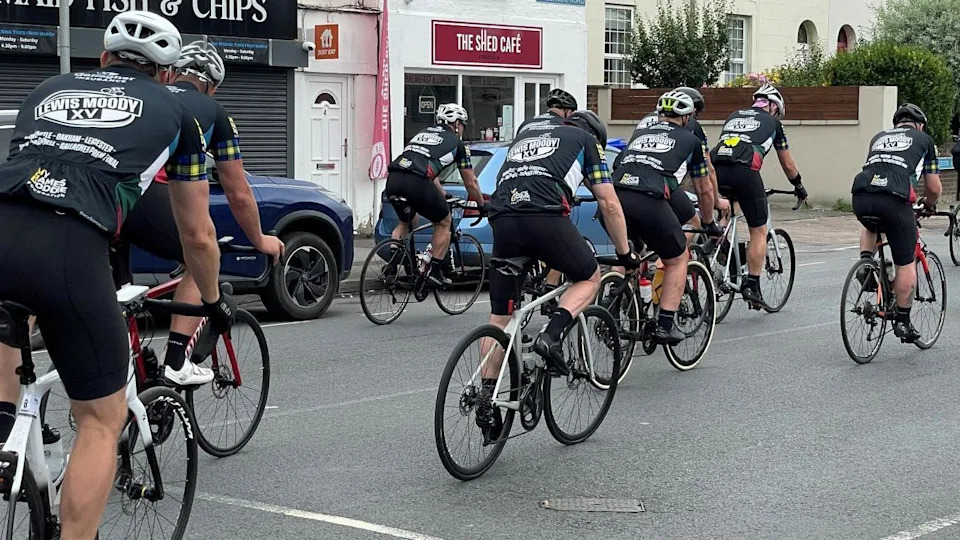 Image of a group of men on bicycles riding on a street lined with cars and shops.