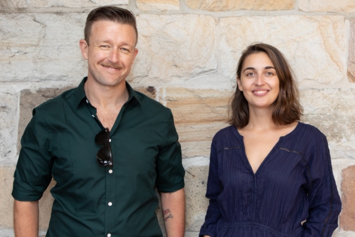 Glenn McGrath and Gabi Billing stand next to each other in front of a stone wall, smiling.