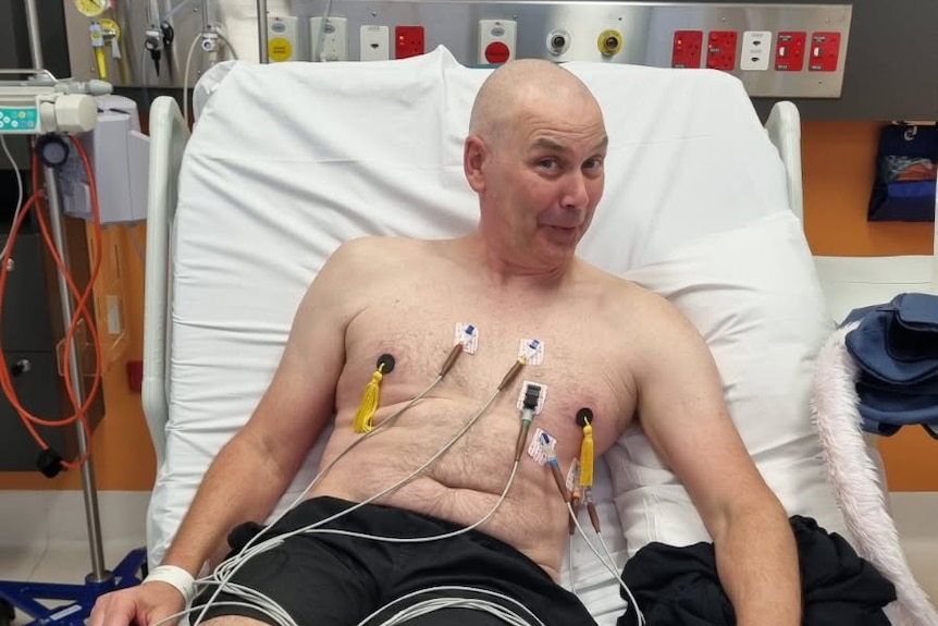 A bald man lying shirtless in a hospital bed, with nipple tassels on.