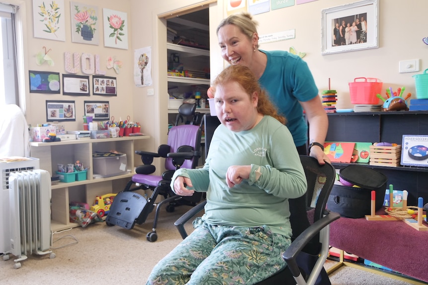 A young woman sitting in a wheelie chair being pushed by another woman inside a room with colourful objects on shelves