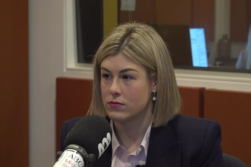 A woman with chin length blonde hair in a dark jacket and white shirt sits behind a microphone with an ABC symbol on it.