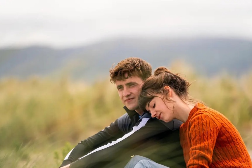 A young couple sits in a field of tall grass, with the woman resting her head on the man's shoulder.