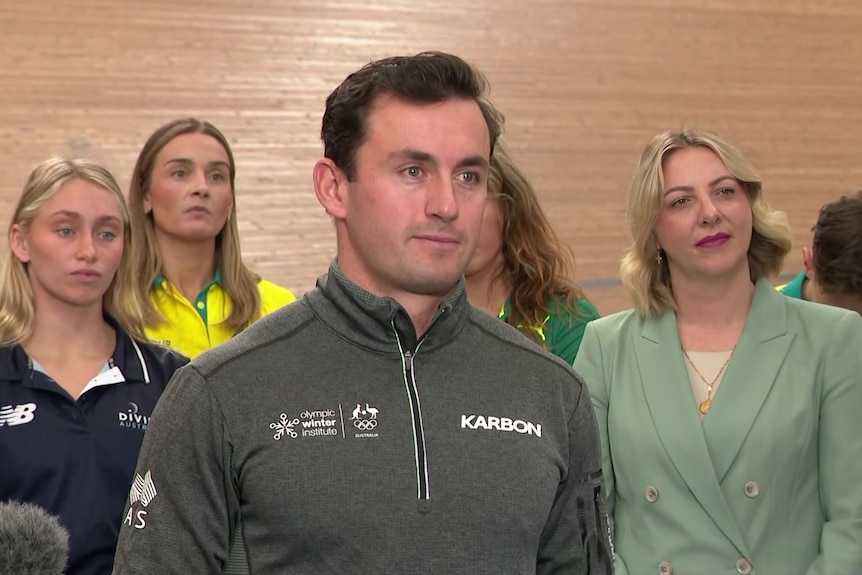 A man in a tracksuit standing in front of wooden wall with women behind him.