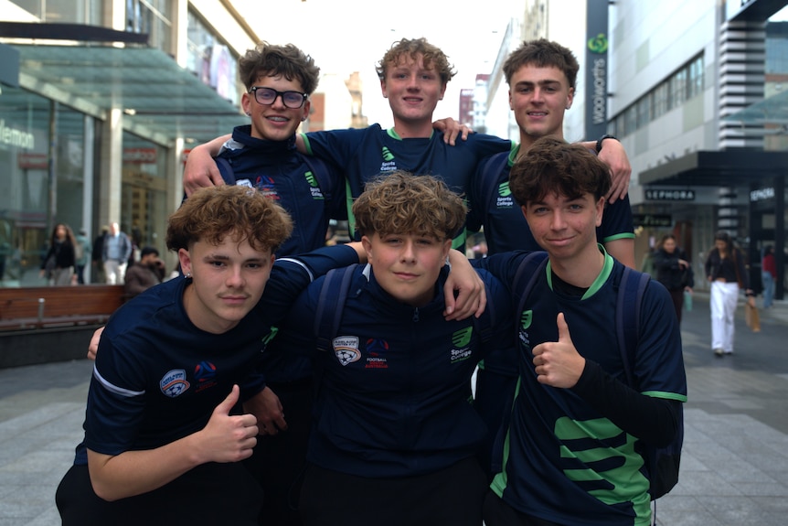 A group of six teenage boys wearing navy and green shirts. Some have put their thumbs up.