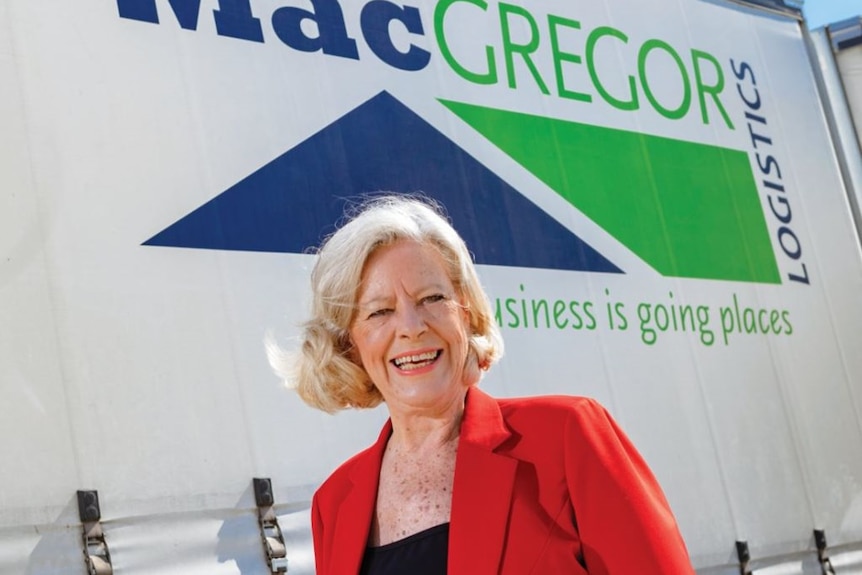 In the background, the side of a truck that reads "Macgregor Logistics". In the foreground a blonde middle-aged woman in red