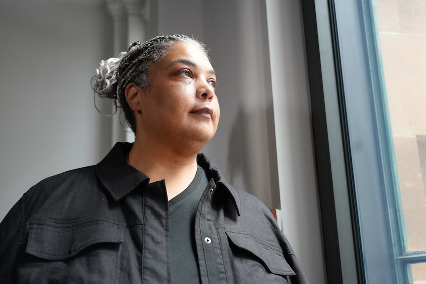 A middle-aged woman with braided hair wears a dark shirt and looks out a window.