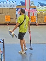 A man in a lime green top and black shorts, with a beard in a hardware store holding a long hammer.
