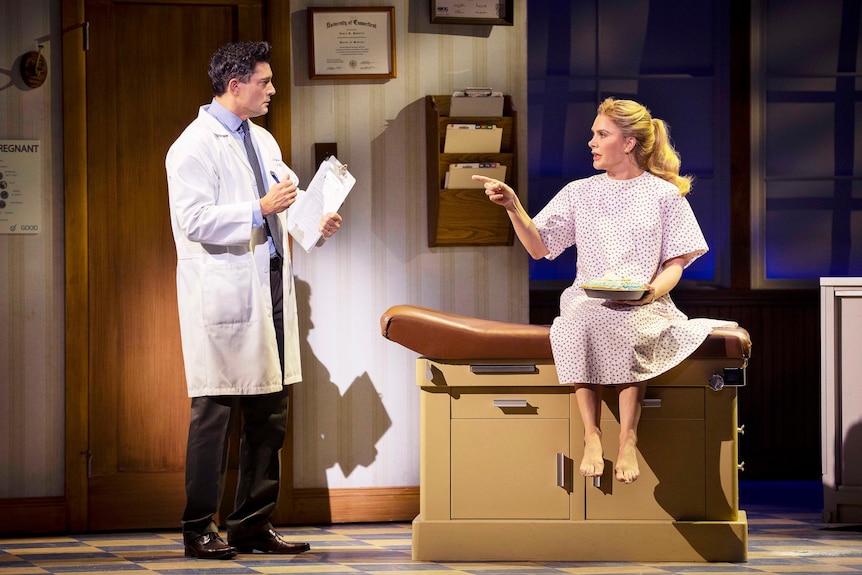 A man stands in a lab coat with a clipboard and speaks to a woman sitting on a hospital bed in a hospital gown.