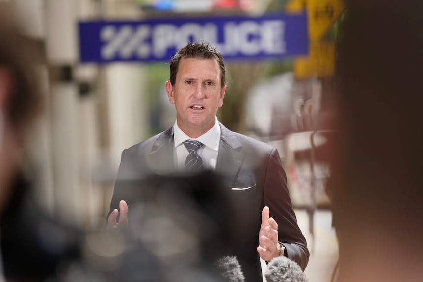 A photo of a man at a press conference. He is wearing a suit ans speaking. Behind him a blurred blue sign says POLICE.