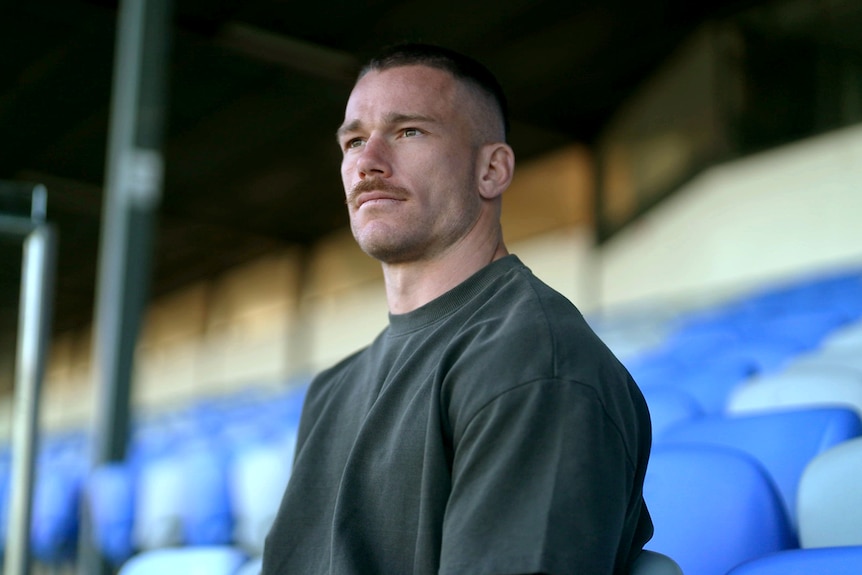 A man wearing a tshirt sits in a grandstand looking ahead with a determined expression.