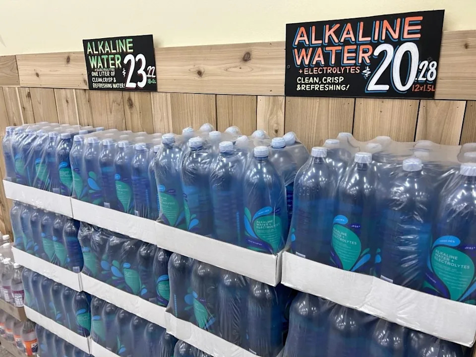 Shelves stocked with alkaline water bottles featuring prices and promotional signs highlighting benefits like electrolytes and refreshment