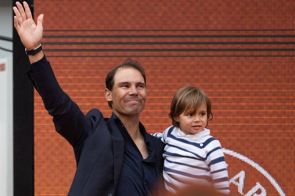 Rafael Nadal in a black shirt and jacket, waving while holding his son Raphael who wears a stripes jumper.