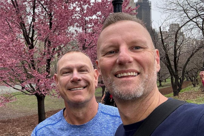 Two men in blue shirts are standing in a garden and smiling at the camera