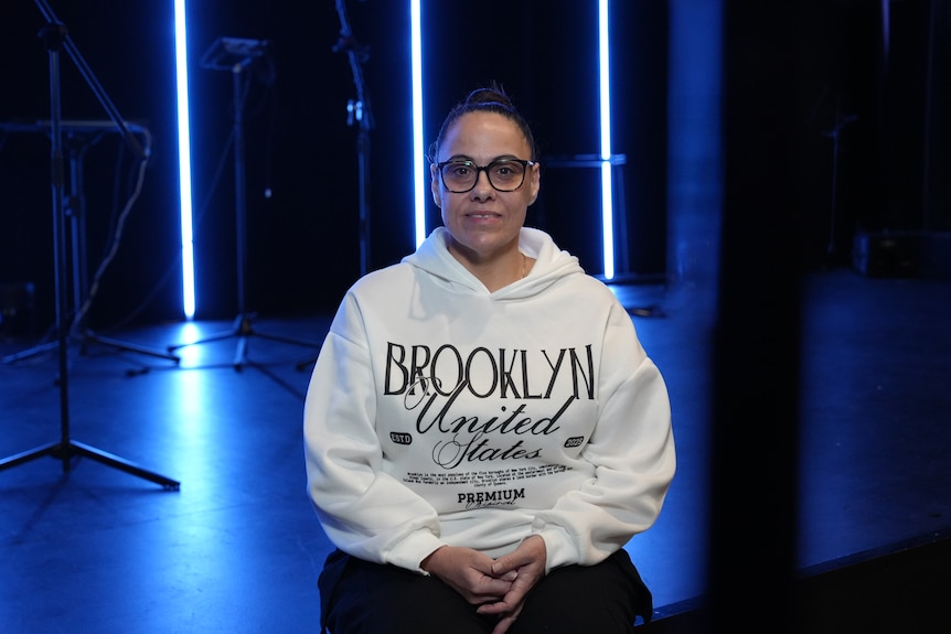 A woman wears a white hoodie as she sits on a stage lit by blue lights