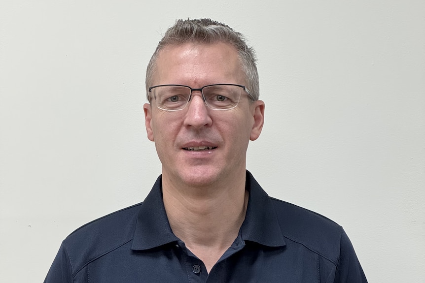 A man in a collared t-shirt stands in front of a light-coloured wall