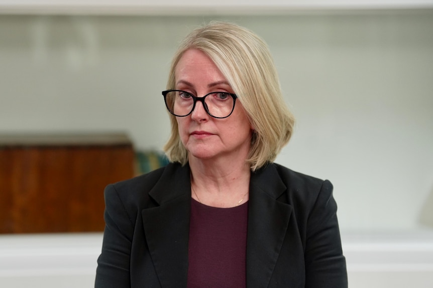 A woman with blonde shoulder-length hair and glasses wearing a black jacket and maroon top