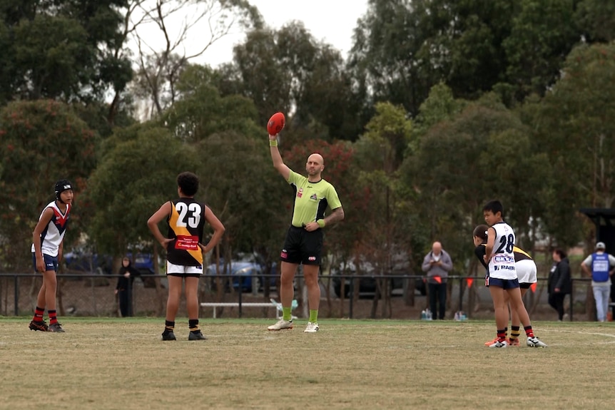Players watch as an umpire holds the ball above his head at a junior Australian Rules football match.