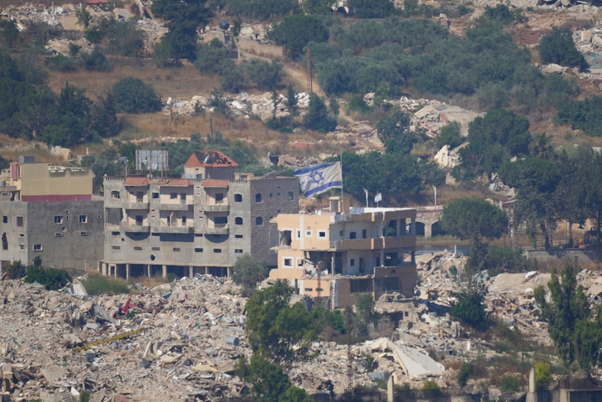A landscape image shows an Israeli flag on top of a multi-storey building that has been bombed, the ground is covered in rubble