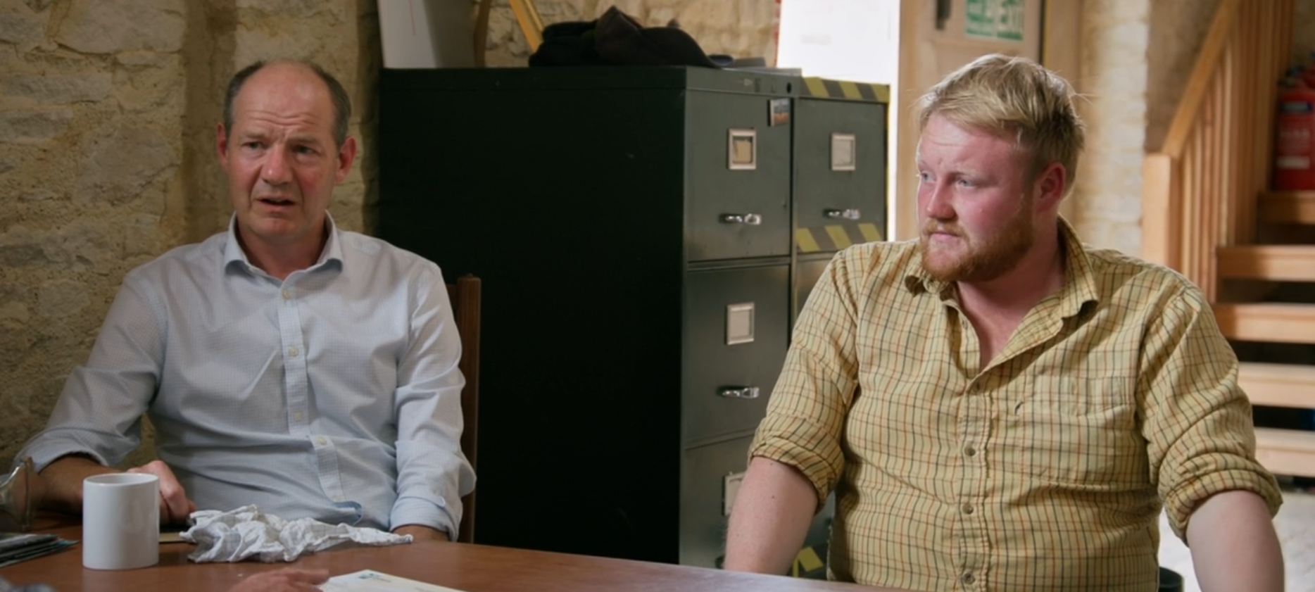 A middle-aged man and a young man look concerned as they sit at a table in a barn.