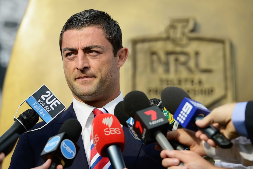 A man with short, dark hair, wearing a suit jacket and tie, speaking while microphones are in front of him.