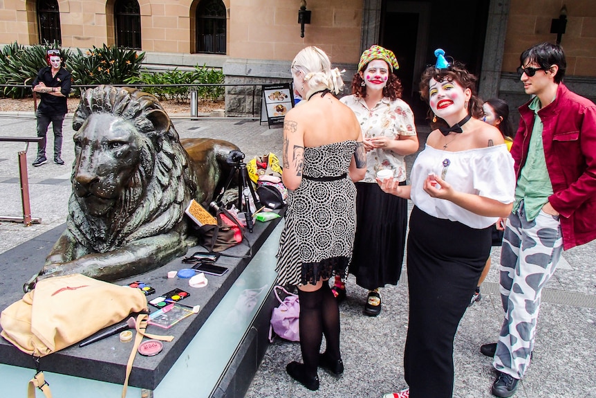 People dressed as clowns stand around a lion statue.