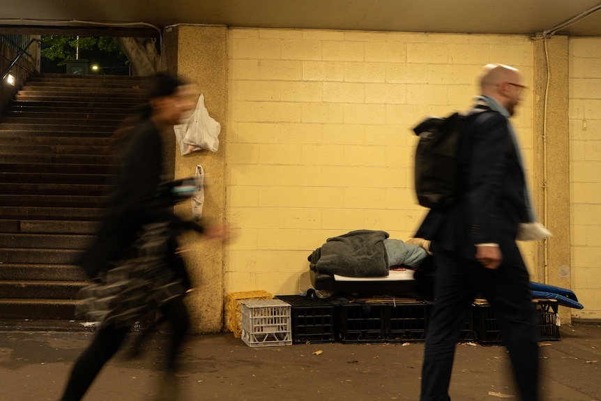 A homeless person lays on a makeshift bed as commuters pass him by.