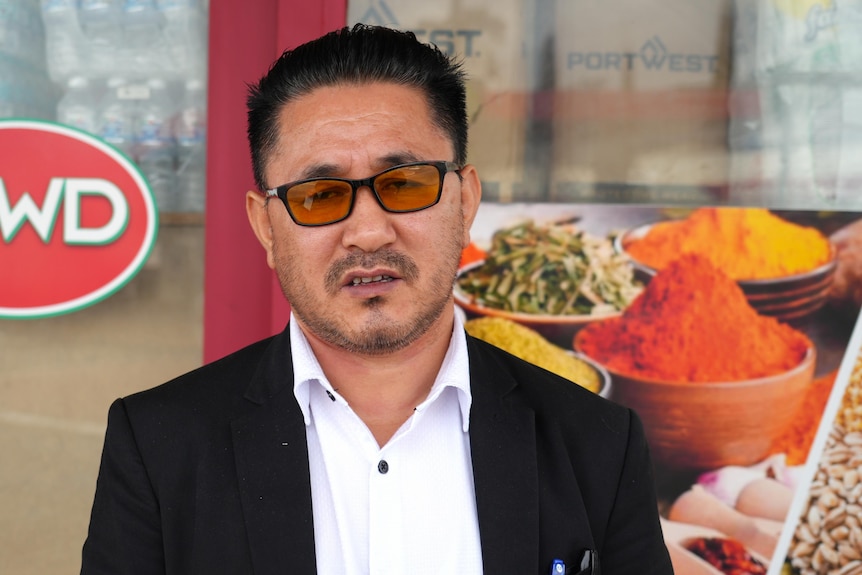 A man wearing a white shirt, black jacket and yellow-lensed glasses stands in front of a door with a red frame.