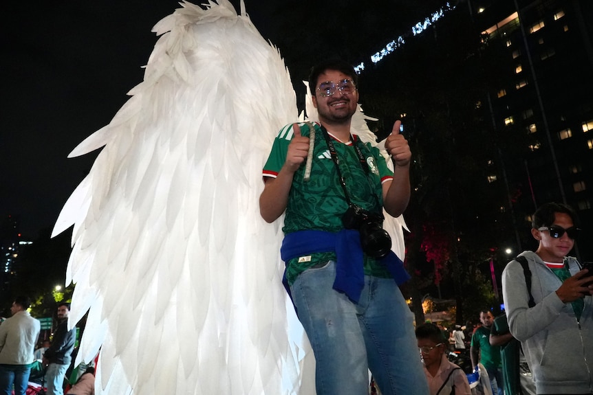 A man wearing a Mexico jersey and giant angel wings gives two thumbs up.