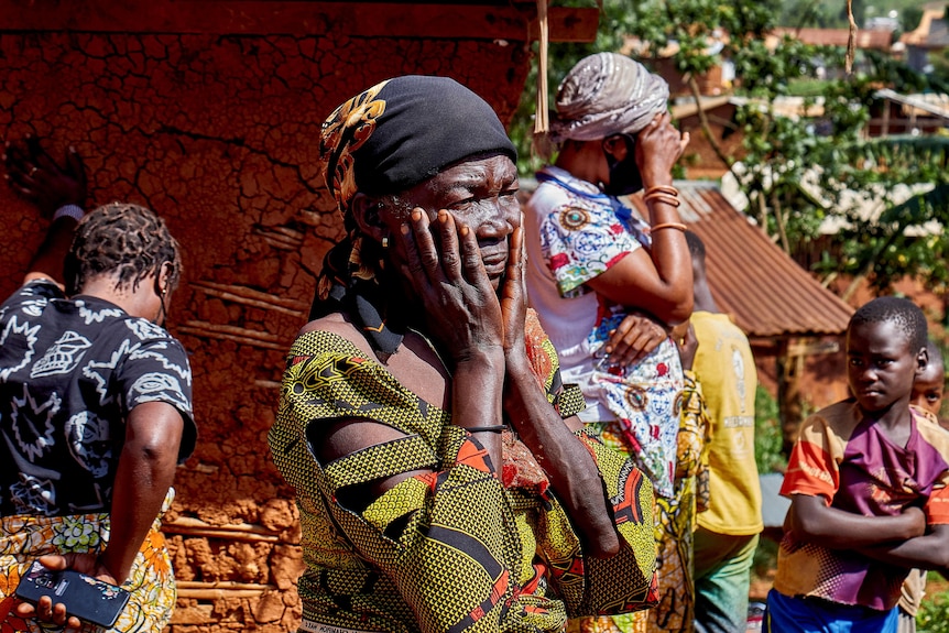 A woman in distress holds her hands to her face as other upset villagers stand around behind her in shock.