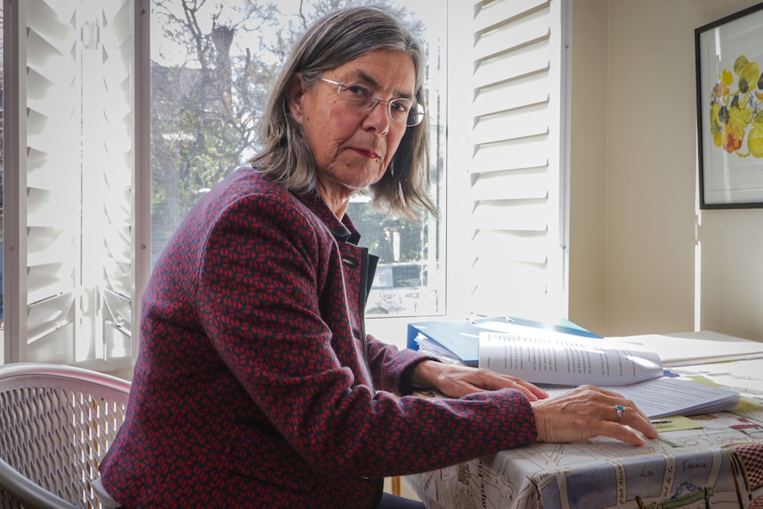 A middle-aged woman with grey hair and glasses sits at a small table with documents on it.