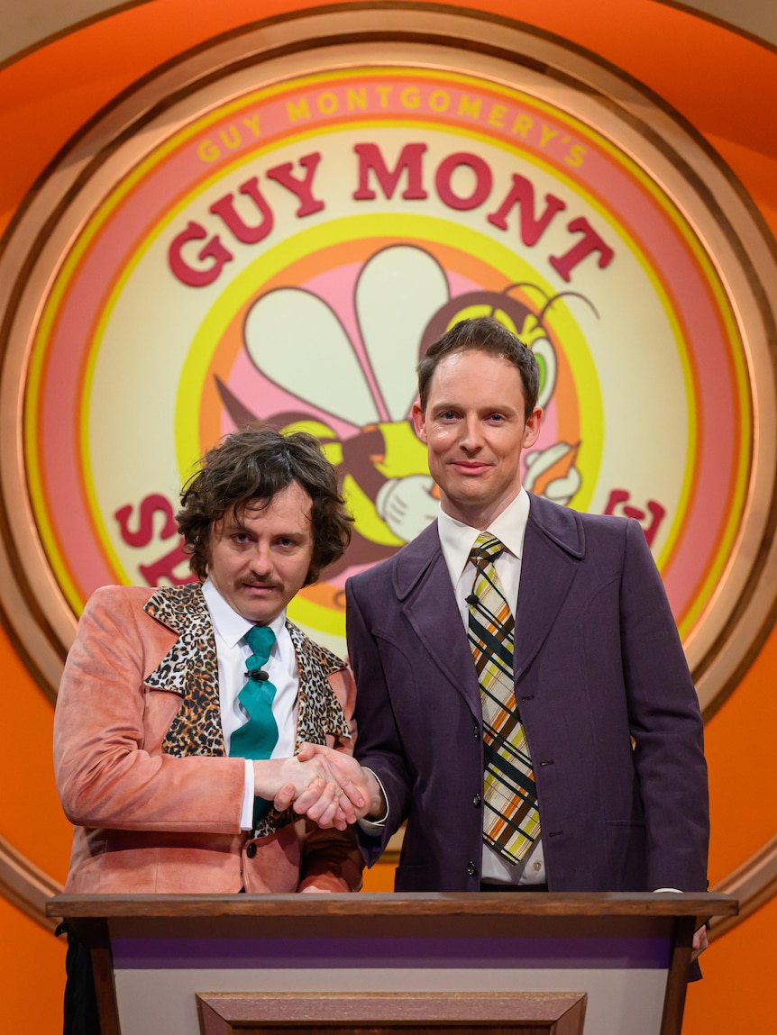 Guy Montgomery in a suit shakes hands with Sam Campbell in a pink jacket with leopard print lapels in front of the show set