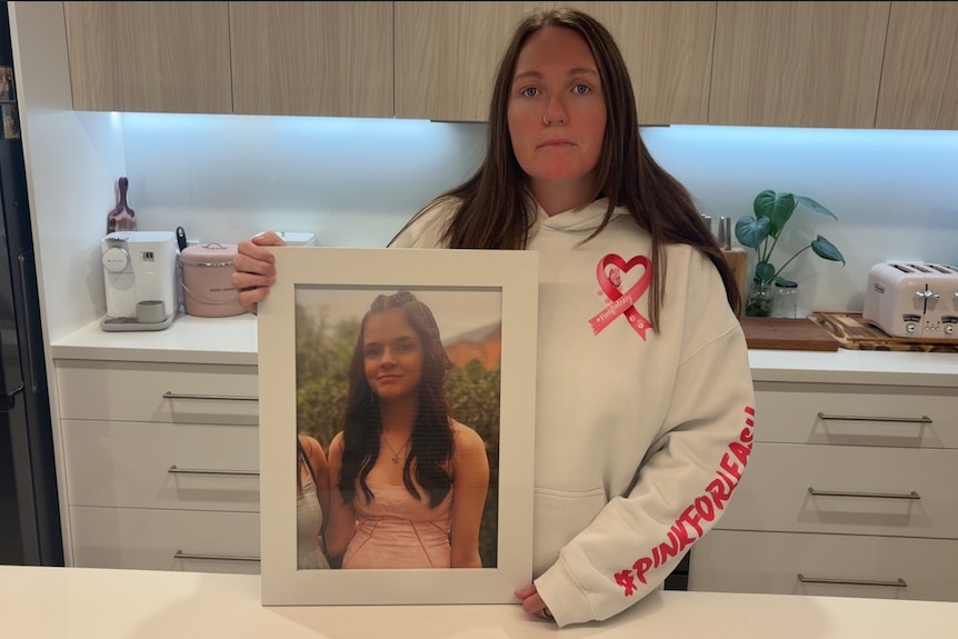 A woman holds a framed photo of Annalyse Blyton. She is wearing a jumper with a pink heart logo and Pink for Leasy written on it