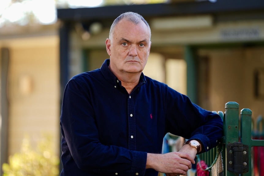 A man with light grey hair stands leaning against a fence with a navy button up shirt.