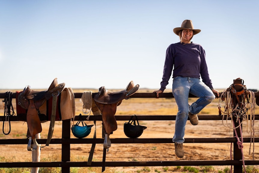 A woman wearing a cowboy hat, sitting on a fence next to horse saddles, smiling at camera