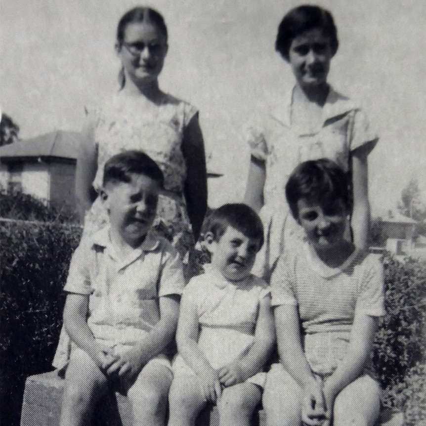 A black and white photo with five kids three of them sitting on a bench with two behind.