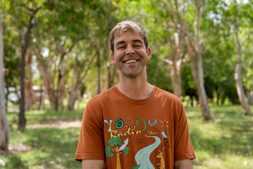Will is wearing a organse shirt that says Yolngu radio. He has blonde and brown wavy hair and is smiling.