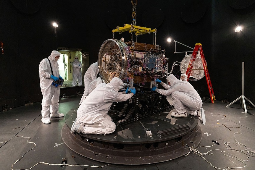 Five people in protective white clothing bending around a bulky machine with a domed ending, in a dark open area.