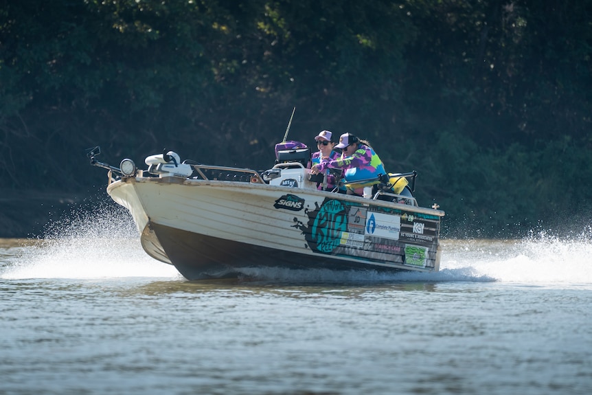 A white small fishing boat cruising along the water, two women in purple / green long sleeve tops driving it.