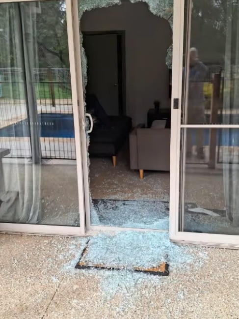 Broken glass covers a door mat and the ground outside a sliding glass door that has been fully smashed.