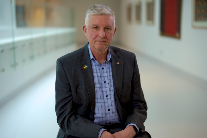 Grant McArthur sitting in a chair in a hospital hallway. Wearing a blue check shirt and suit