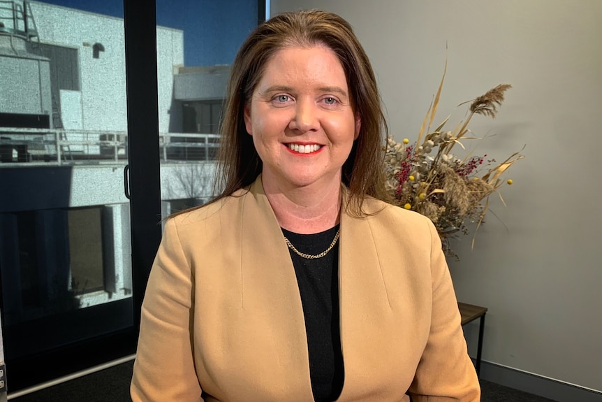 Liz smiling, Caucasian, wears beige jacket, black tee, dried flowers behind, glass doors, blue sky, building visible.